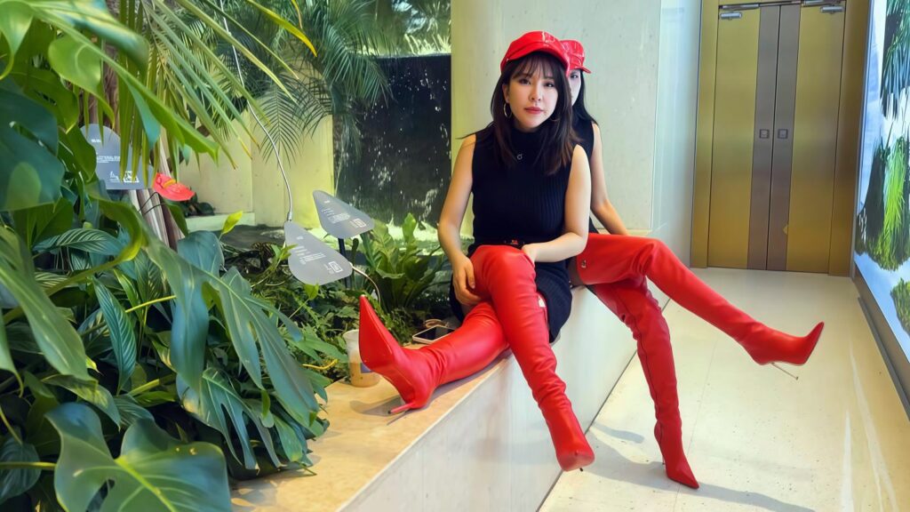 D25BTV034 - Ein Mobiltelefon, zwei Paar leuchtend rote Overknee-Stiefel mit starkem Gefühl der Umhüllung und eine spielerische Revolution unter einer V-Zeichen-Geste.